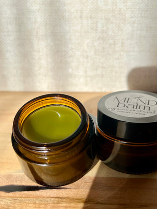 Open jar of comfrey infused jojoba and tallow balm with a closed jar labeled 'Mend Balm' on a wooden surface.