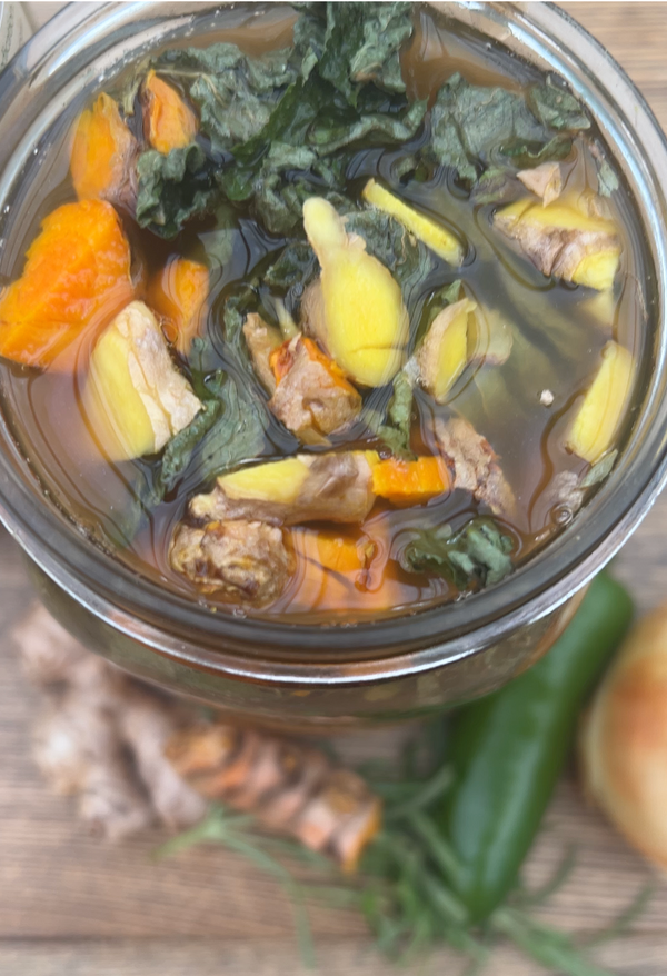 A close-up overhead view of a glass jar filled with a clear broth containing kale leaves, ginger slices, orange carrot pieces, and chunks of meat, with additional ginger and a green chili blurred in the foreground on a wooden surface.