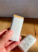 A herbal balm stick of organic muscle joint balm for stiff joints and sore muscles, showing the beige balm stick end, with the matching white cap lying on a wooden surface in the background.
