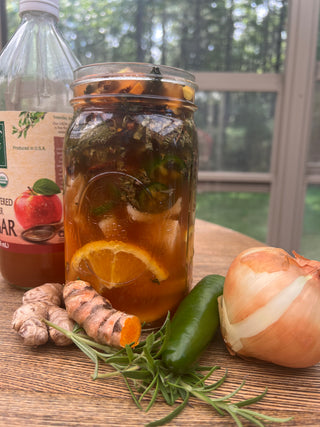A large glass mason jar filled with iced tea, garnished with an orange slice, resting on a wooden table with ingredients including ginger, a jalapeño, rosemary, and an onion. A bottle of apple cider vinegar is visible in the background.