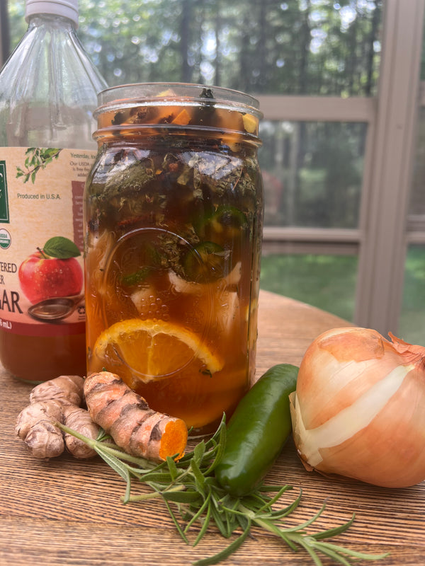 A large glass mason jar filled with iced tea, garnished with an orange slice, resting on a wooden table with ingredients including ginger, a jalapeño, rosemary, and an onion. A bottle of apple cider vinegar is visible in the background.