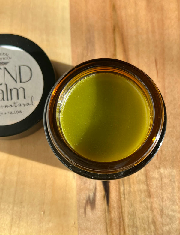 Open jar of green balm on a wooden surface with a lid labeled 'Balm'. Comfrey infused jojoba oil and tallow balm