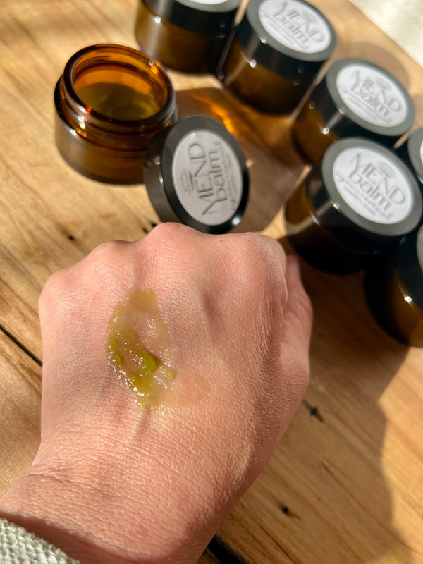 Hand with green comfrey and tallow balm on wrist next to bottles labeled 'Mend Balm' on a wooden surface