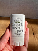 A hand holding a white stick of Natural Northern Organic Muscle + Joint Balm for stiff joints and sore muscles against a wooden table and textured gray wall background.