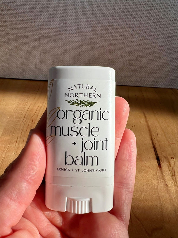 A hand holding a white stick of Natural Northern Organic Muscle + Joint Balm for stiff joints and sore muscles against a wooden table and textured gray wall background.