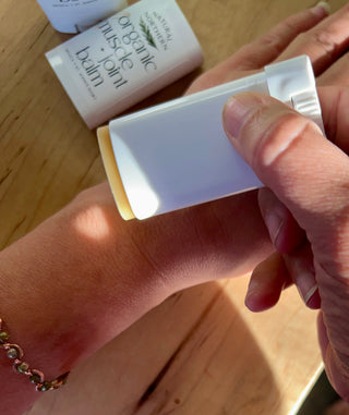 applying a white herbal balm for stiff joints and sore muscles stick to their wrist, with two white containers of Nature's Nutrition Organic Muscle Balm visible in the background on a wooden surface.