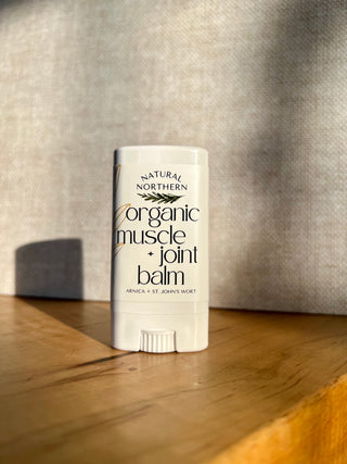 A white stick of herbal balm labeled 'Natural Northern Organic Muscle + Joint Balm'  for stiff joints and sore muscles, made with arnica and st.john's wort. 