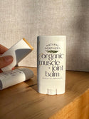 A stick of Natural Northern Organic Muscle + Joint Balm stands on a wooden surface, with a hand holding another herbal balm in the background against a textured beige wall.