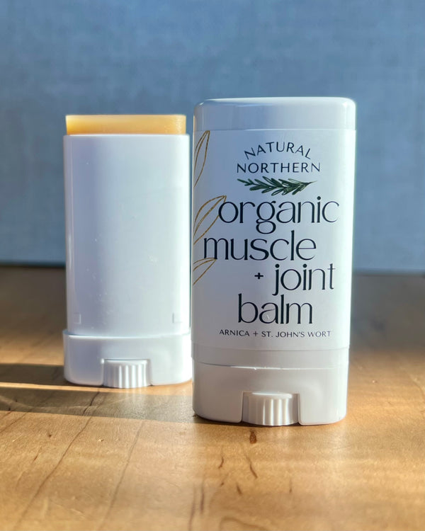 Two white Natural Northern Organic Muscle + Joint Balm herbal balm sticks on a wooden surface against a blue-gray background. One stick is open, showing a pale yellow balm stick top, and the other is closed with printed branding visible.