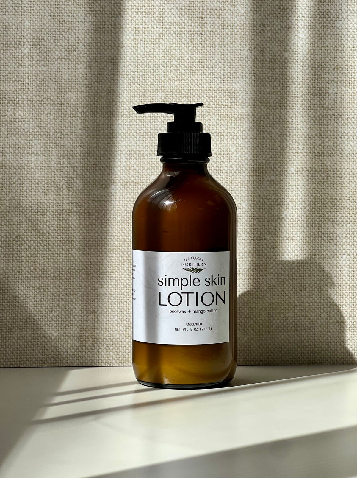 An amber glass pump bottle of Simple Skin Lotion for sensitive skin,  a black pump and white label, set on a white surface against a textured beige curtain with soft window shadows. 